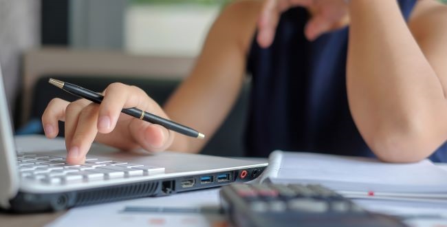 A kép illusztráció, melyen irodai munkakörnyezetben egy alkalmazott keze látható, amint számítógépen és számológépen dolgozik.