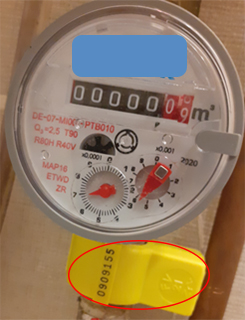 A képen egy vízmérő óra látható, mely alatt egy sárga műanyag biztonsági záróelem látható.