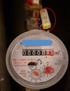 A képen egy vízmérőóra látható, mely felett zsinórral rögzítve van egy fehér-sárga biztonsági záróelem.
