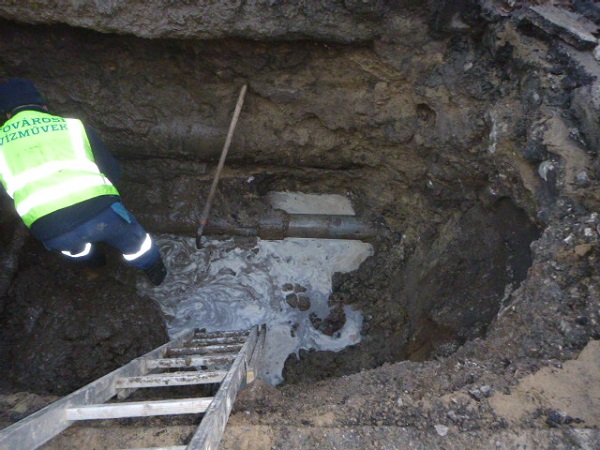 A képen egy mély gödör alján dolgozó csővezetéket cserélő munkás látható. 