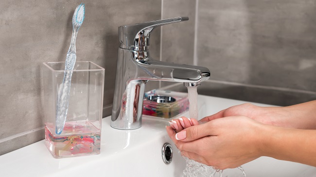 A képen egy mosdó, egy pohárban tárolt fogkefe, valamint egy kéz látható kézmosás közben.