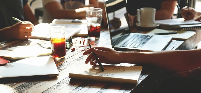 A képen egy papírokkal és laptoppal ellátott asztal, három emberrel, akikből csak a kar látható. Dolgoznak éppen.